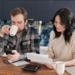 Casal analisa contas domésticas com calculadora e celular em uma cozinha.