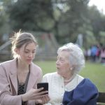 Mulher orienta uma idosa no uso do celular em um parque.