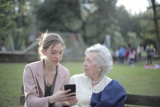 Mulher orienta uma idosa no uso do celular em um parque.