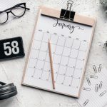 Calendário sobre uma prancheta, acompanhado de lápis, artigos de papelaria e óculos sobre uma mesa.