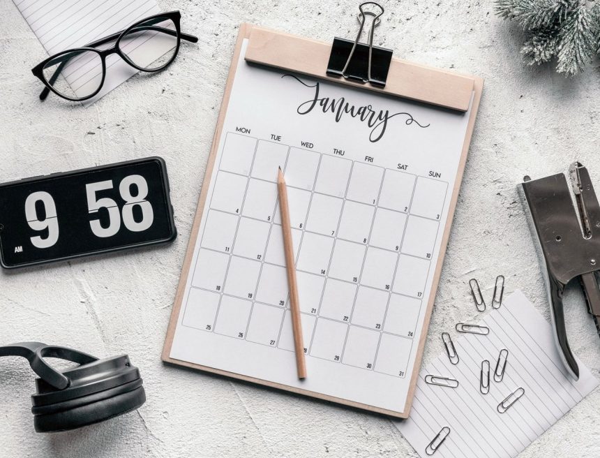 Calendário sobre uma prancheta, acompanhado de lápis, artigos de papelaria e óculos sobre uma mesa.